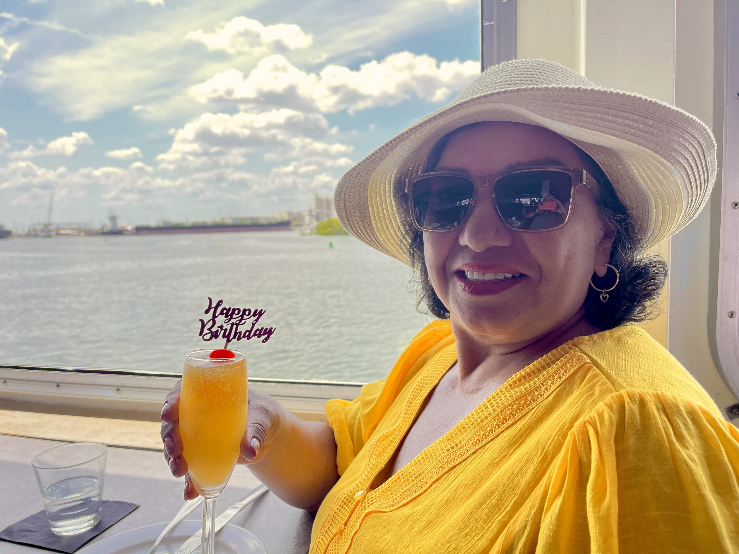 Ana Magalhaes enjoying a cocktail adorned with a happy birthday decoration, with a beautiful beach view in the background through a window